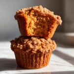Two stacked sweet potato muffins, one cut in half showing the moist interior and crumb topping.