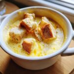 Close-up of a white bowl filled with bubbling yellow cheese fondue topped with toasted bread cubes.