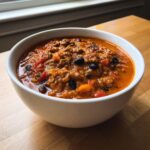 A close-up of a white bowl filled with rich, thick pumpkin chili featuring ground meat and black beans.