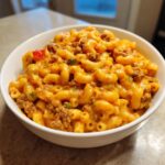 A close-up of a white bowl filled with creamy, cheesy taco pasta mixed with ground beef and small pieces of peppers.
