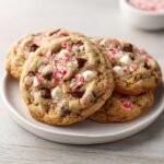 Four freshly baked peppermint chocolate chip cookies loaded with white chocolate chips and crushed candy canes on a light plate.