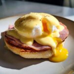 Close-up of a classic Eggs Benedict with runny yolk spilling out from the poached egg under rich hollandaise sauce.