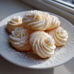 A plate of freshly baked, piped butter cookie swirls heavily dusted with powdered sugar, catching sunlight.