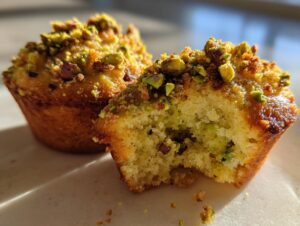 Close-up of a golden-brown pistachio muffin, broken open to show the moist interior and topped with chopped pistachios.
