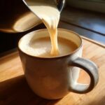 Close-up of creamy white hot chocolate being poured from a metal pot into a speckled ceramic mug.