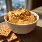 A white bowl filled with bright orange pumpkin fluff dip, served next to a stack of square crackers.