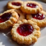 A close-up of several golden, buttery raspberry thumbprint cookies with bright red jam centers arranged on a white plate.