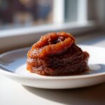 A close-up of a dollop of rich, dark brown pear butter served on a small white plate, highlighted by natural light.