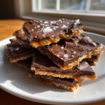 A stack of broken pieces of saltine cracker toffee, showing the caramel, chocolate, and flaky sea salt topping.