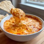 A tortilla chip scoops a generous portion of hot, cheesy chili cheese dip from a white bowl.