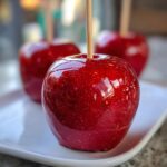 Close-up of a perfectly coated, shiny red candy apple with a wooden stick, sitting on a white plate.