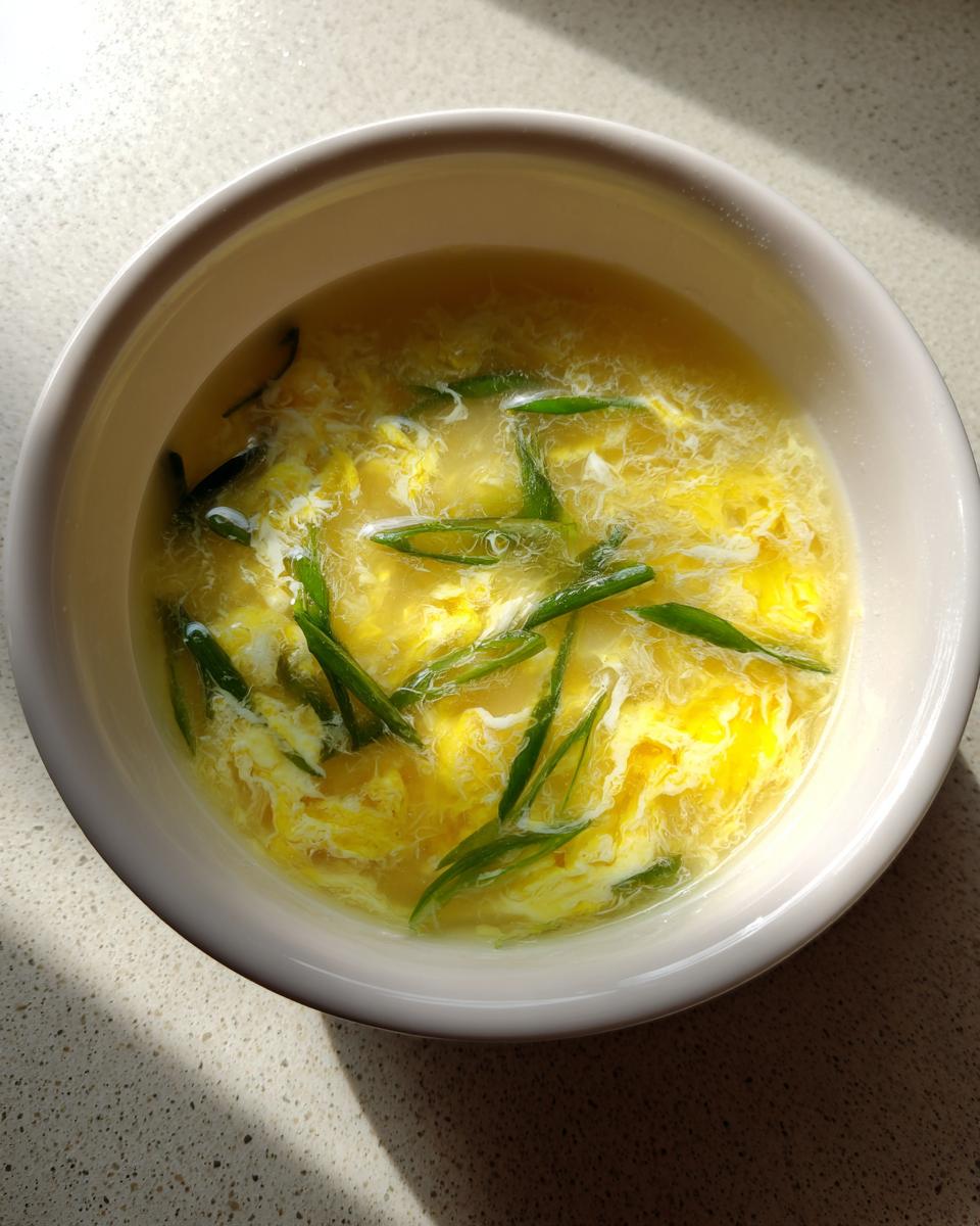 Overhead view of a bowl of classic egg drop soup, featuring wispy yellow egg ribbons and bright green scallions.