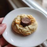 A single, freshly baked peanut butter cup cookies resting on a small white plate, held by a hand.