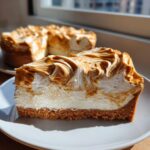 A close-up slice of creamy biscoff cheesecake featuring a thick graham cracker crust and a dramatic swirl of white and caramelized biscoff topping.