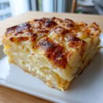 A close-up of a thick, square slice of potatoes au gratin showing layered potatoes and creamy sauce, topped with golden brown cheese.