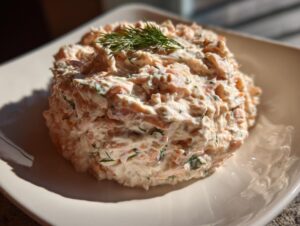 A mound of creamy smoked salmon dip garnished with a sprig of fresh dill, served on a white square plate.