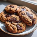 Close-up of several chewy sourdough chocolate chip cookies with melted chocolate chunks on a light-colored plate.