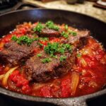 Close-up of seared steak pizzaiola pieces simmering in a rich, red tomato and onion sauce, topped with fresh parsley.