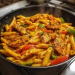 Close-up of steaming Rasta Pasta tossed in a creamy, orange sauce with seasoned chicken pieces and colorful bell peppers.