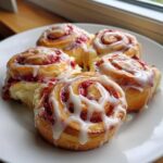 Four delicious Strawberry Cheesecake Sweet Rolls drizzled generously with white cream cheese icing on a white plate.