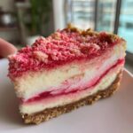 A close-up slice of strawberry crunch cheesecake featuring a graham cracker crust and bright pink strawberry crunch topping.