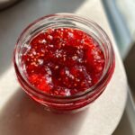 Close-up overhead view of vibrant red strawberry freezer jam with visible seeds in a small glass jar.
