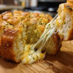 A piece of hot cheese bread is pulled away, showing long, gooey strings of melted cheese stretching from the loaf.