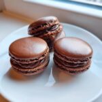 Three rich, dark chocolate macarons with ruffled feet and creamy filling stacked on a white plate.
