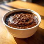 Close-up of thick, dark brown pureed black bean soup served in a white bowl on a wooden table.