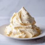 A large, perfectly piped mound of creamy, pale yellow vanilla buttercream frosting on a white plate.