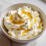 A close-up of fluffy whipped goat cheese topped with golden honey and a sprinkle of pepper.