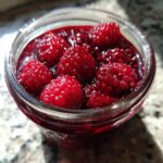 Close-up of whole, bright red raspberries suspended in thick, glistening raspberry jam inside a glass jar.