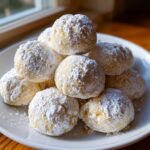 A mound of soft, round lemon cookies heavily coated in white powdered sugar on a white plate.