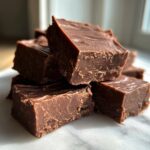 A stack of rich, dark chocolate fudge squares resting on a white marble surface, highlighting the smooth texture of the fudge.