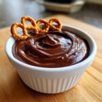 A close-up of a creamy, rich chocolate dip in a white ramekin, garnished with three small pretzels.