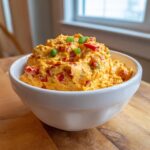 A close-up of creamy, orange boat dip mixed with red peppers and topped with green onions in a white bowl.