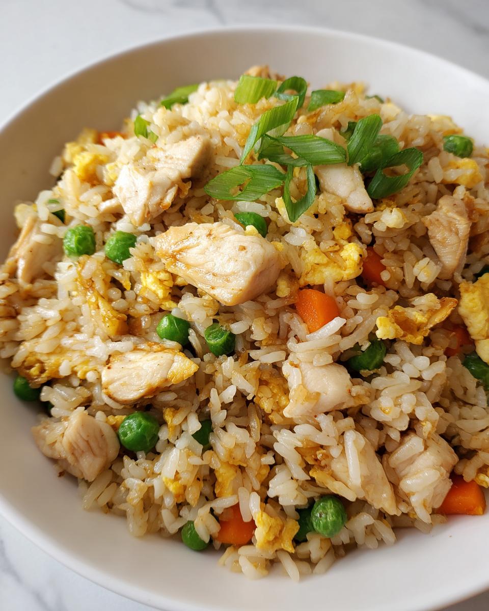 A close-up of a white bowl filled with amazing chicken fried rice, mixed with scrambled egg, peas, carrots, and topped with green onions.