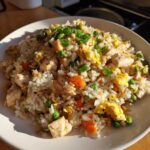 A close-up of a serving of amazing chicken fried rice, featuring white rice, diced chicken, carrots, peas, egg, and green onions.