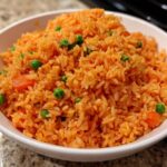 A close-up of a white bowl filled with vibrant orange Mexican rice mixed with bright green peas and diced carrots.