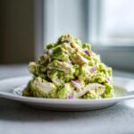 A mound of creamy avocado chicken salad mixed with shredded chicken and diced red onion served on a white plate.
