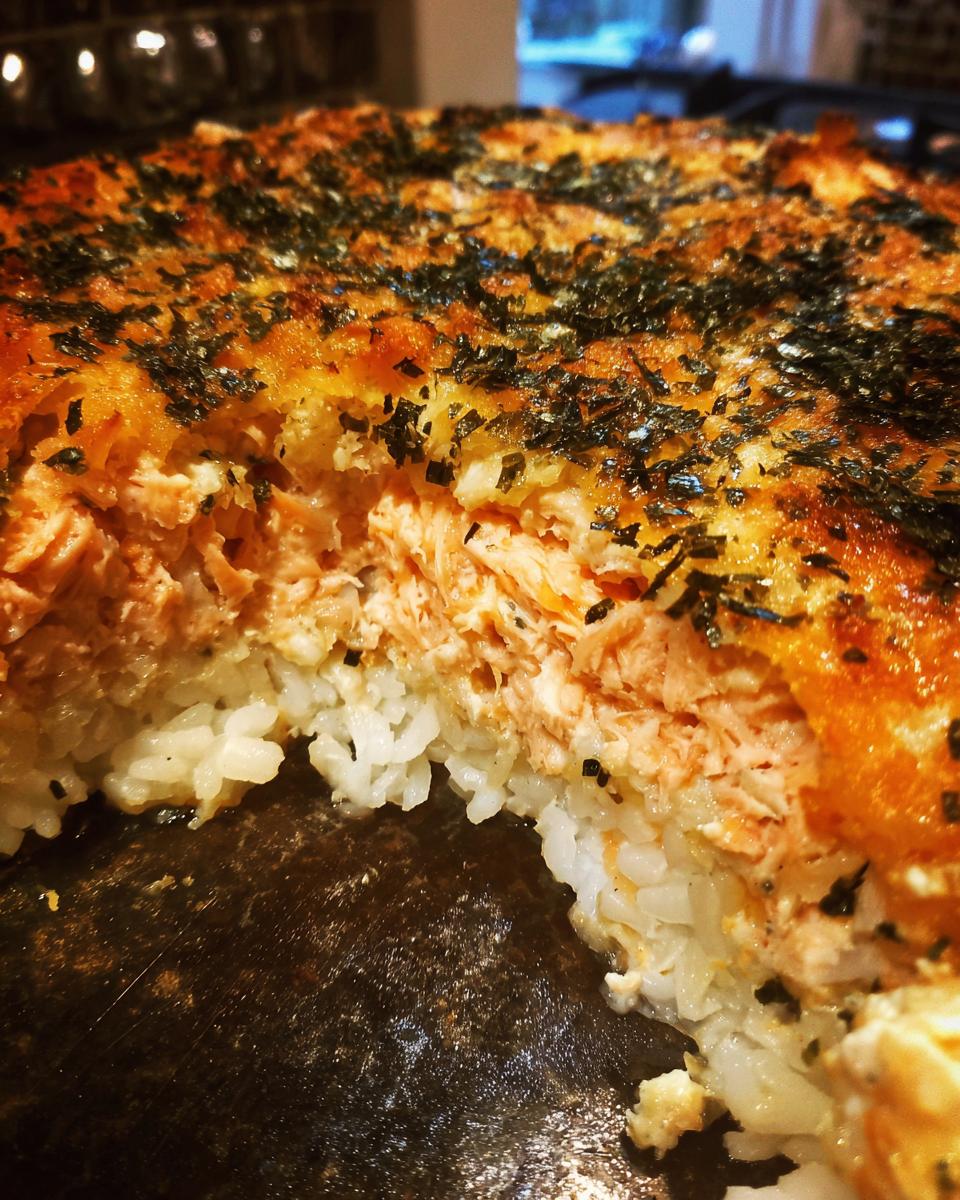 A close-up cross-section of a baked sushi bake showing layers of rice, creamy salmon filling, and a browned, seaweed-topped crust.