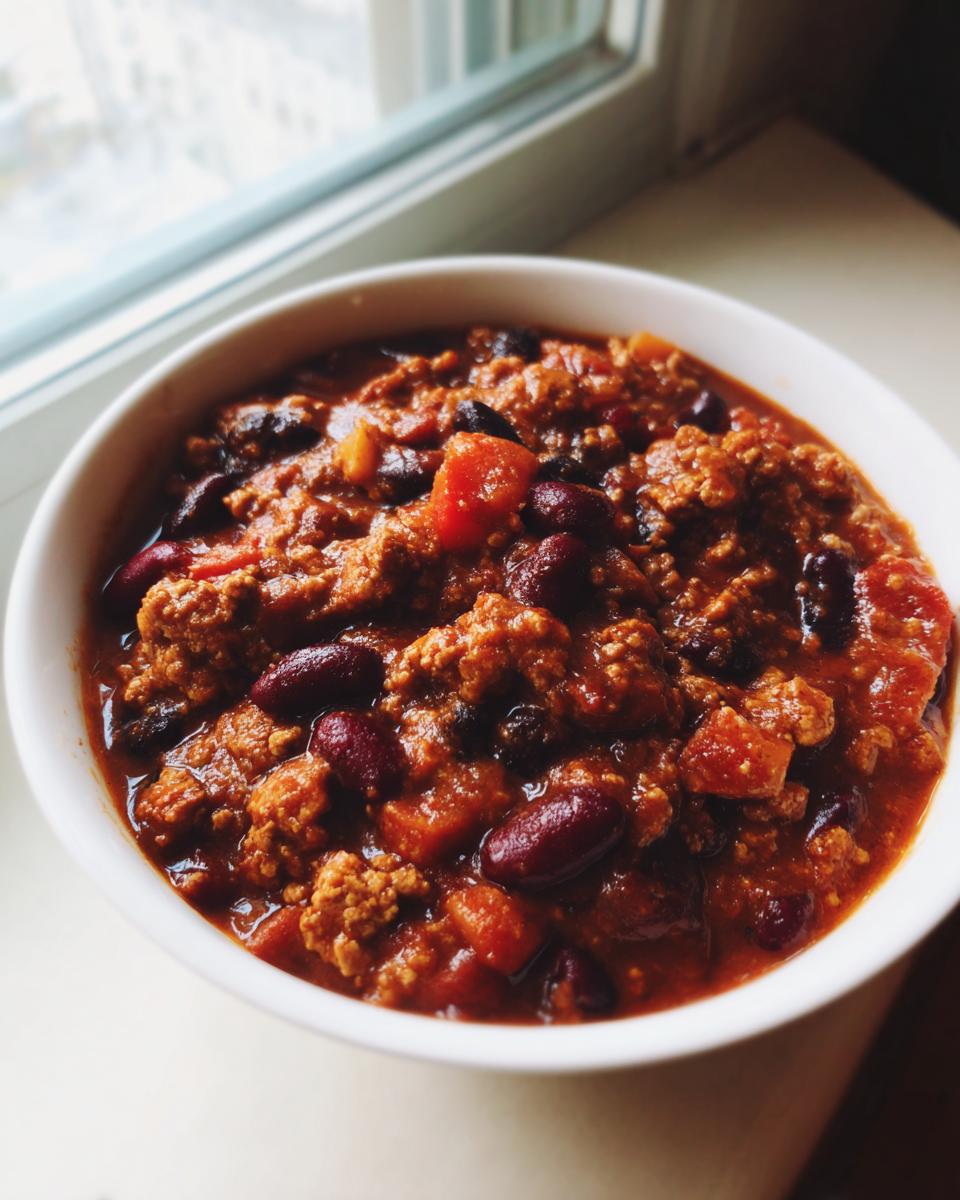 A close-up of a white bowl filled with rich, thick high protein chili featuring ground meat and kidney beans, set near a bright window.