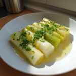 Four segments of tender braised leeks served on a white plate, glistening in butter sauce and topped with fresh parsley.