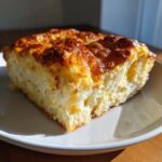 A square slice of fluffy, cheesy egg bake with a deeply browned, bubbly top, served on a white plate.
