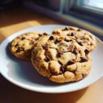 Close-up of three thick, chewy chocolate chip cookies piled on a white plate, highlighted by natural window light.