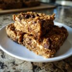 Three chewy oatmeal bars stacked on a white plate, featuring visible oats and chocolate chips.