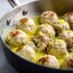 Close-up of browned chicken piccata meatballs simmering in a bright yellow lemon-butter sauce, garnished with zest.