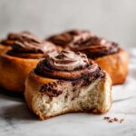 Close-up of a soft, fluffy chocolate cinnamon rolls with a bite taken out, topped with rich chocolate frosting.