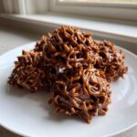 A small pile of glossy, chocolate-coated haystack cookies resting on a white plate.