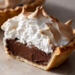A cross-section of a delicious chocolate meringue pie showing the rich dark filling and fluffy, toasted meringue topping.
